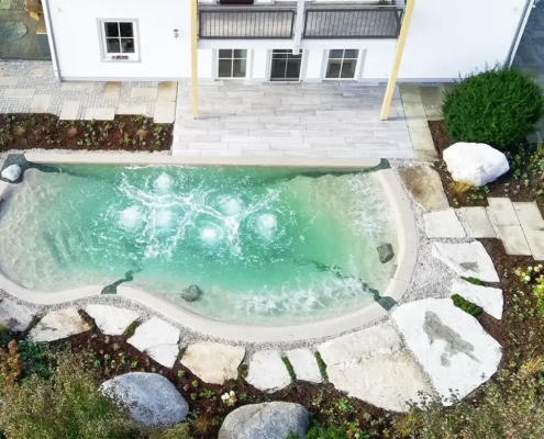 Bio.design-Pool in geschwungener Form vor Terrasse mit Randbepflanzung und Gartendusche