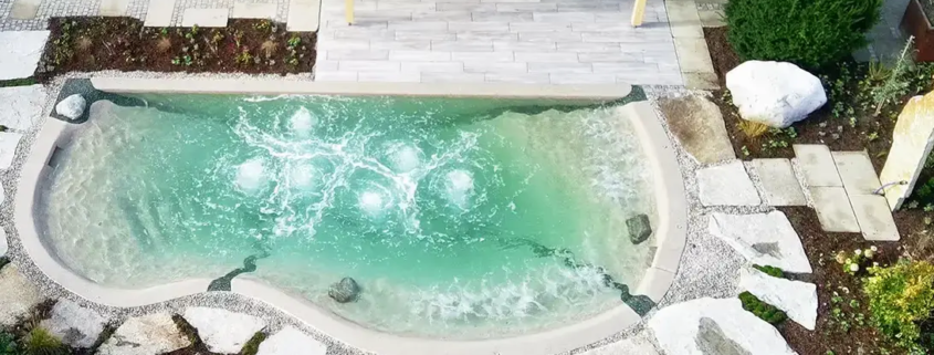 Bio.design-Pool in geschwungener Form vor Terrasse mit Randbepflanzung und Gartendusche