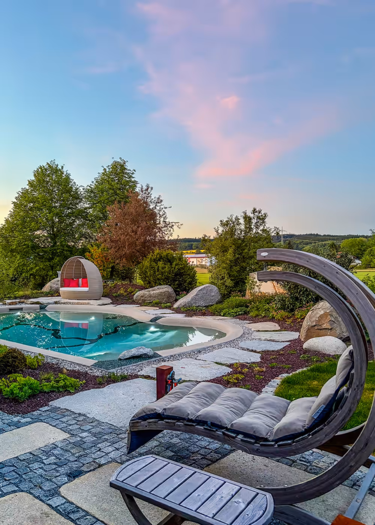 Abendstimmung am Bio.design-Pool: Schaukelsessel im Vordergrund, kokonförmiges Daybed im Hintergrund, einladendes Ambiente für laue Sommerabende
