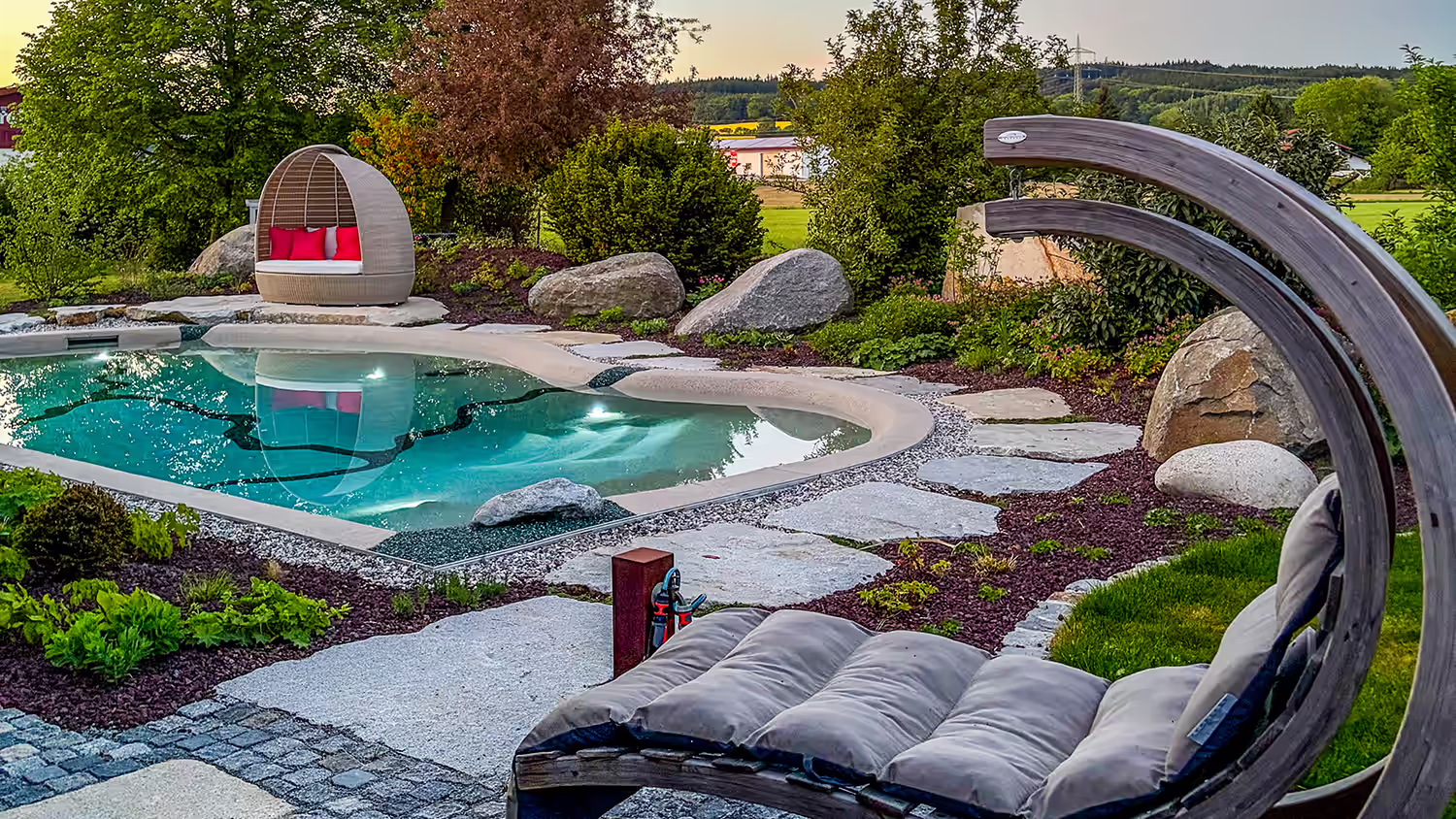 Abendstimmung am Bio.design-Pool: Schaukelsessel im Vordergrund, kokonförmiges Daybed im Hintergrund, einladendes Ambiente für laue Sommerabende