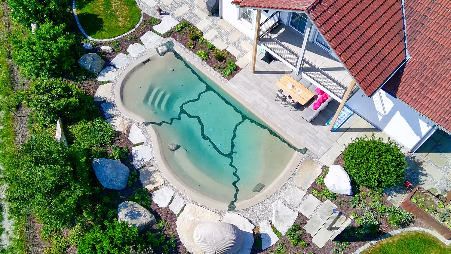 Vogelperspektive auf einen naturnahen Designer-Pool mit angrenzender Terrasse und harmonisch gestalteter Gartenanlage